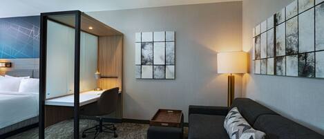 A hotel room with a bed, desk, chair, couch, and artwork on the walls.