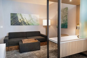 Moderni hotellihuone, jossa on harmaa sohva, puinen sivupöytä, suuri peili ja aaltoa kuvaava taulu.