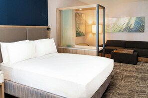 Hotellihuone, jossa on suuri sänky, oleskelualue sohvineen ja pienenä pöytineen, lasiseinä ja taulu seinällä.