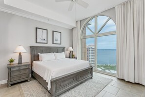 A bedroom with a large bed, two bedside tables with lamps, a view of the sea, and a cityscape through a large window.