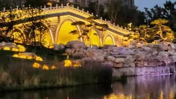 A stone bridge with arches, illuminated by warm lighting, over a body of water with reflections.