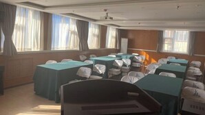 Un salón de conferencias con mesas y sillas, ventanas amplias con cortinas y un proyector en el techo.