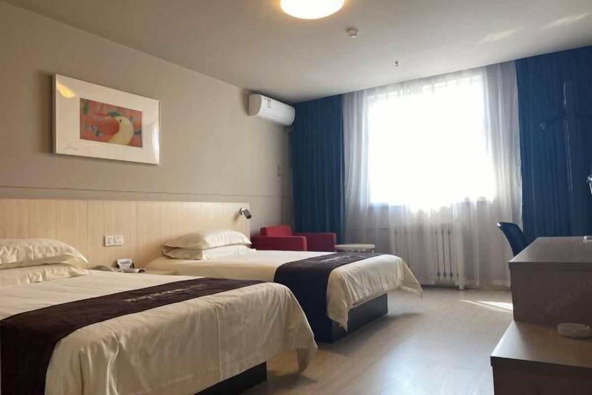 A hotel room with two beds, a desk, a chair, a window with curtains, an air conditioning unit, and a framed picture on the wall.
