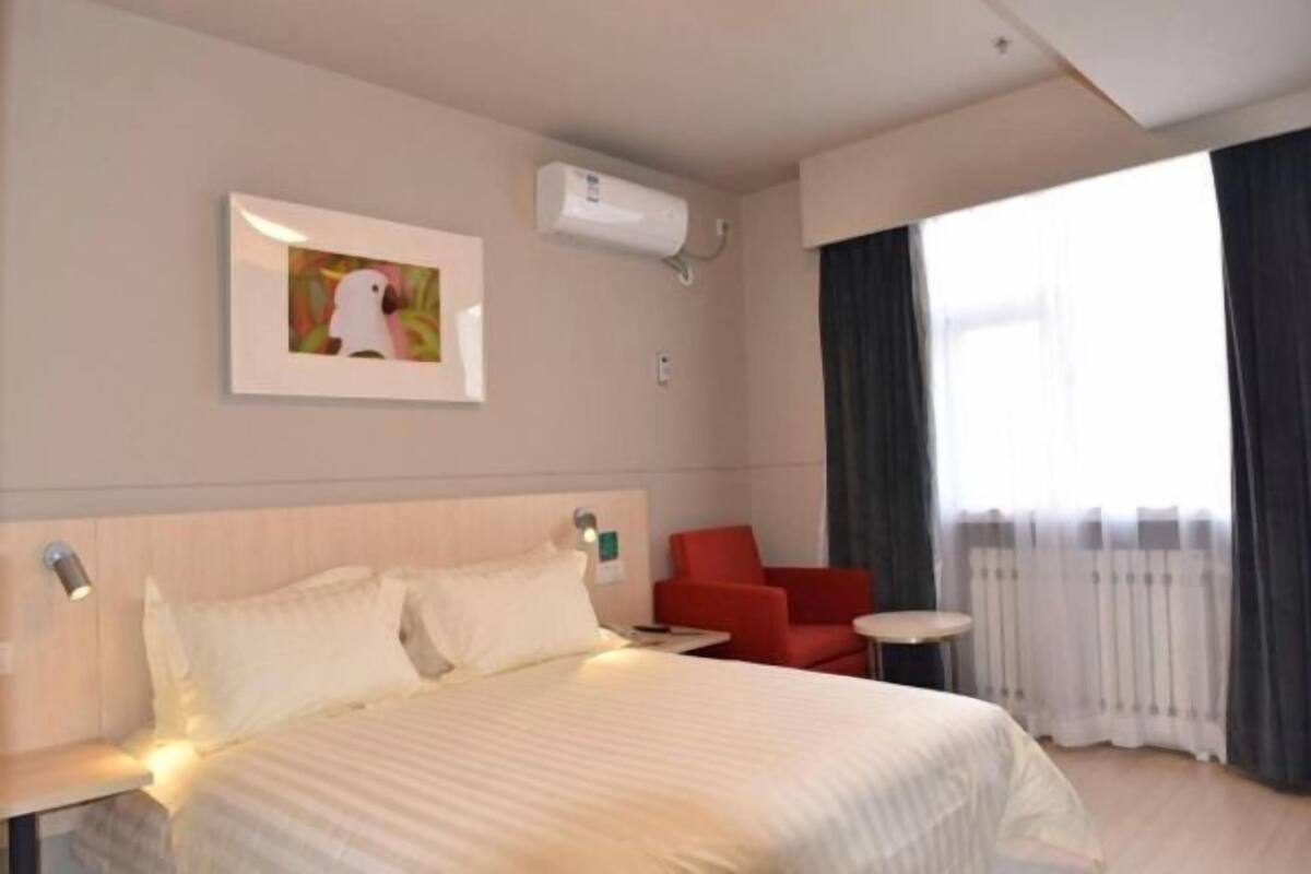 A hotel room with a bed, a red chair, a small table, a wall-mounted air conditioner, and a window with curtains.