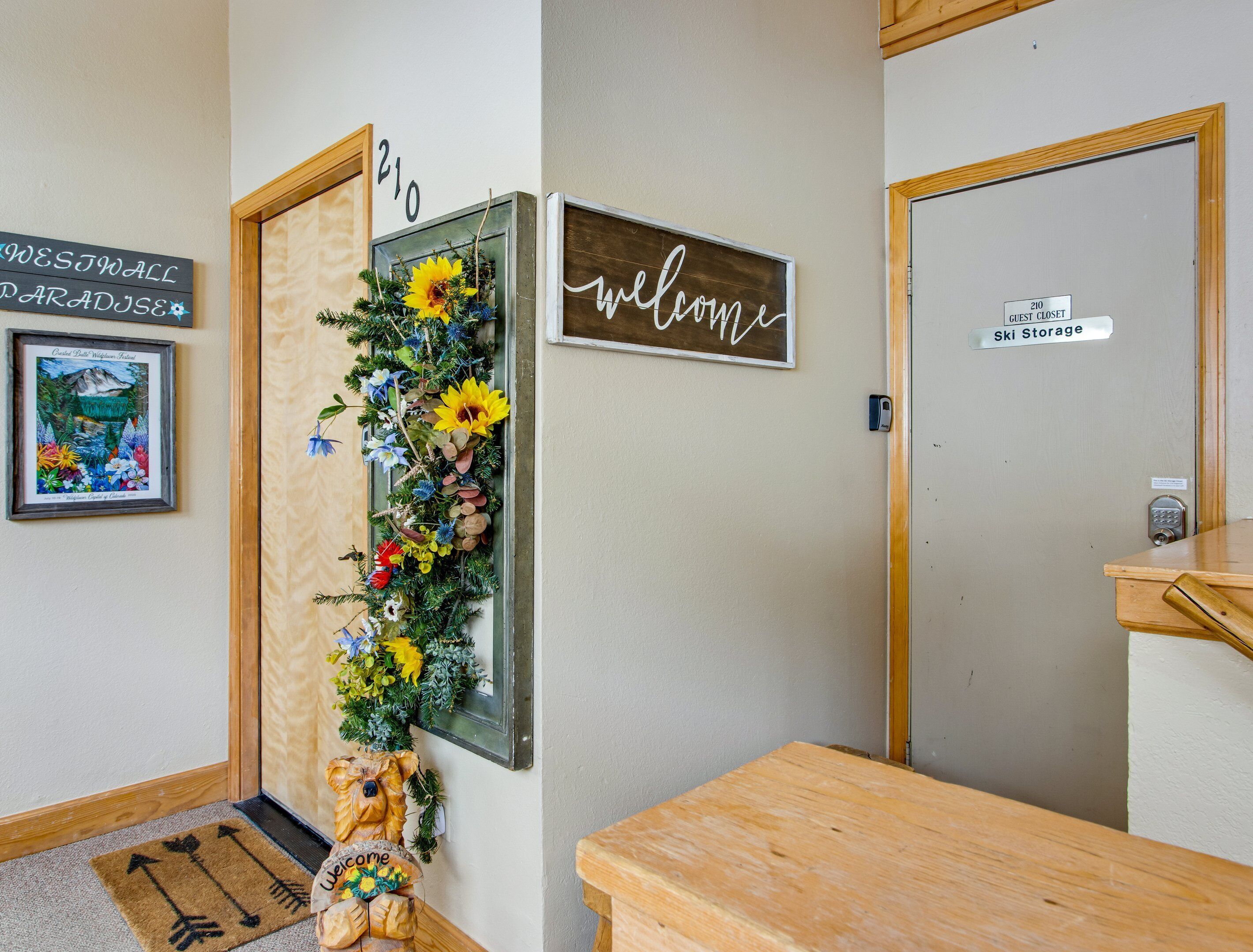 Un espace d’accueil d’hôtel avec un comptoir de réception en bois, un panneau d’accueil, un tableau encadré, un panneau mural portant un numéro et une porte indiquant « Local à skis ».