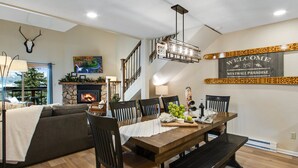 Un coin repas avec une table en bois, des chaises et un banc. On y trouve une cheminée avec un manteau en pierre, un escalier et une pancarte indiquant : « Bienvenue au paradis de Westwall ».