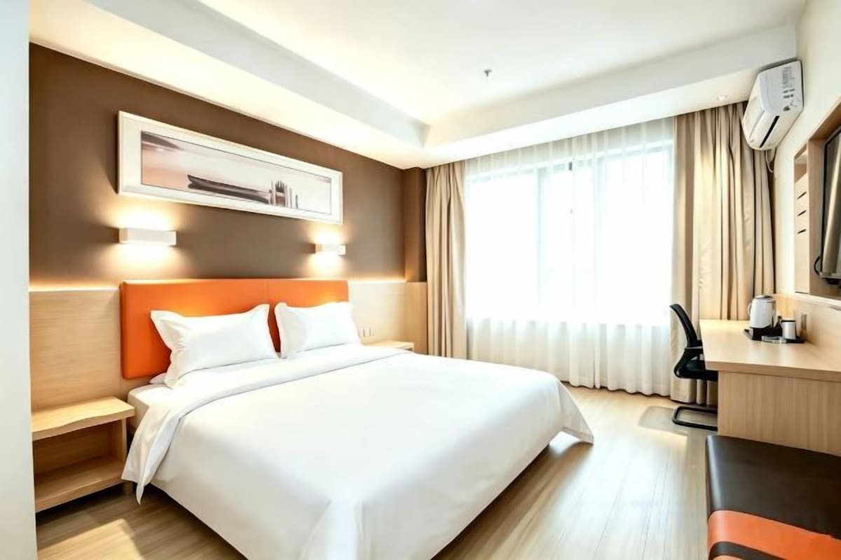 A hotel room with a bed, bedside table, desk, and a window with curtains.