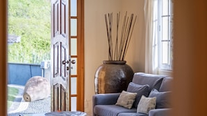 A room with a sofa, a large vase with sticks, a door, and a view of greenery outside.