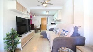Een moderne woonkamer met een flatscreen televisie, een grijze bank, een plafondventilator en een kleine keuken op de achtergrond.