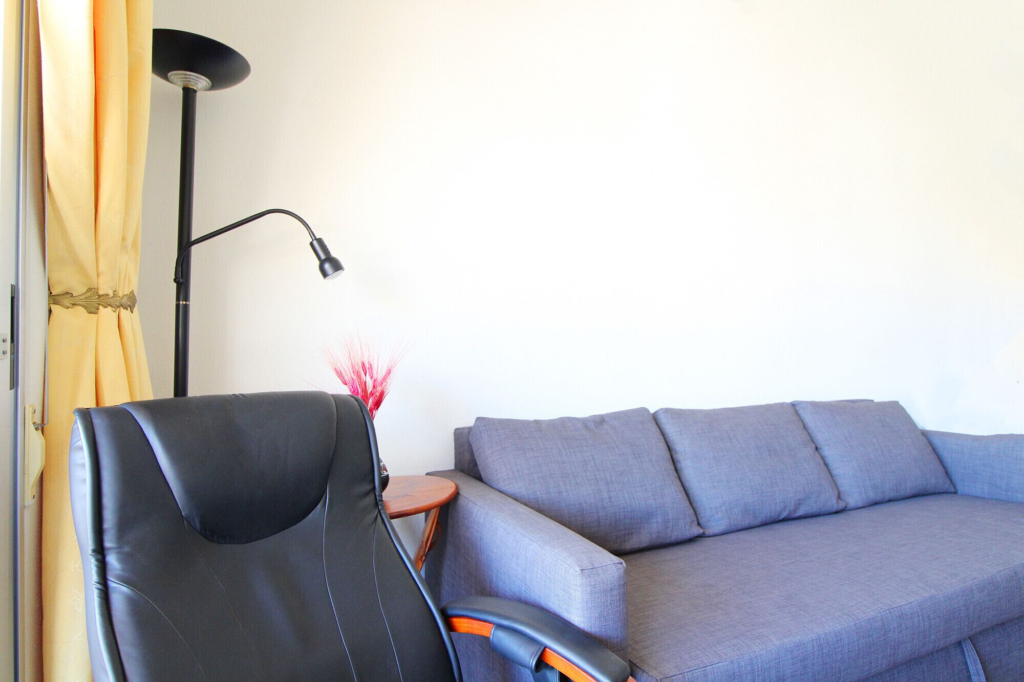 A modern living room with a grey sofa, a black leather chair, a floor lamp, and a small table with a vase of red flowers.