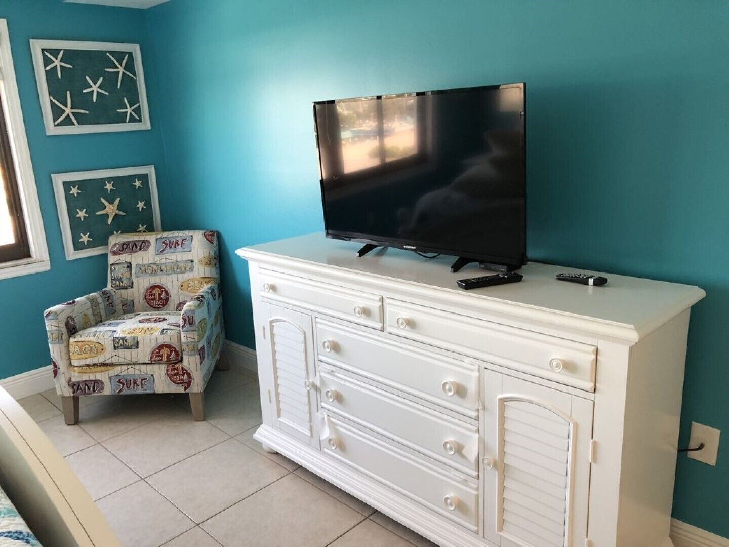 A room with a teal wall, a white dresser with drawers, a television on top, and a patterned armchair.