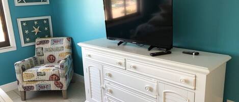A room with a teal wall, a white dresser with drawers, a television on top, and a patterned armchair.