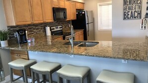 A modern kitchen with granite countertops, stainless steel appliances, and wooden cabinets. There are three bar stools with cushioned seats. A decorative sign on the wall reads "MEMORIES ARE MADE AT THE BEACH." - SANDCASTLES OCEAN VIEW TOP FLOOR..DATES AVAILABLE (check Reviews) (Cocoa Beach)