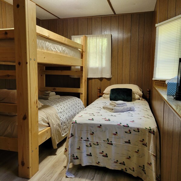 Ein Schlafraum mit Etagenbett, Holzboden, einem Einzelbett mit gemustertem Bezug und einem Fenster mit Jalousien.