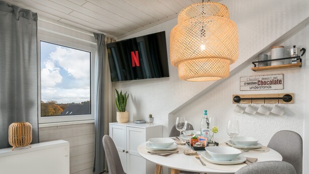 Uma sala de jantar moderna com uma mesa redonda, cadeiras cinzas, uma TV montada acima e vista do céu pela janela.