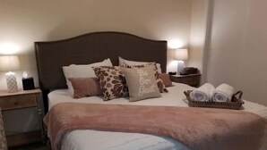 Una cama bien hecha con un cabecero marrón, varios almohadones y una cesta con toallas enrolladas a un lado.