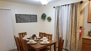 Un comedor con una mesa de madera dispuesta para seis personas, rodeada de sillas de madera. Hay una planta decorativa en la pared, un extintor de incendios montado y una ventana con cortinas.