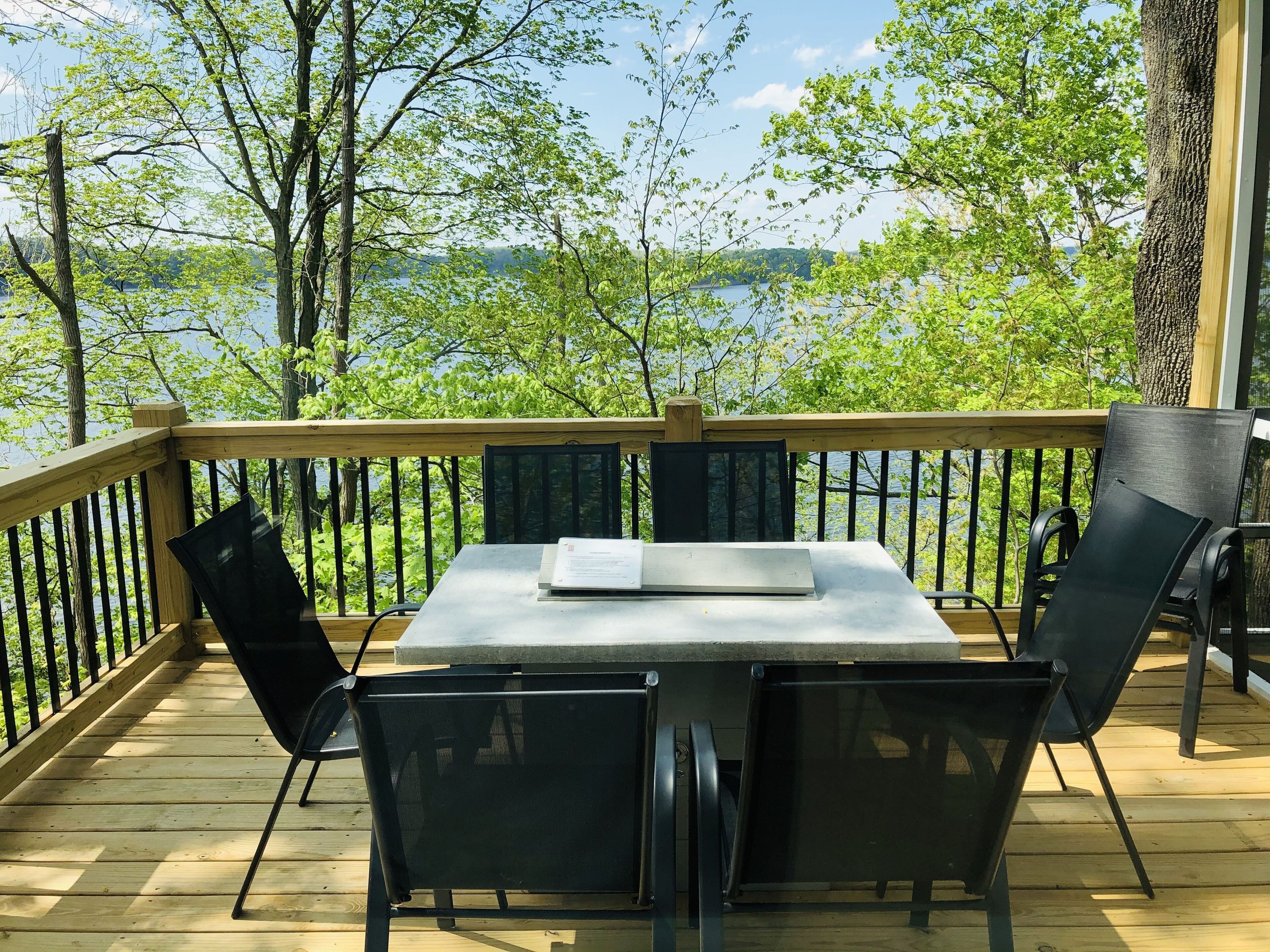 A wooden deck with a table and chairs, overlooking a lake with trees in the background.