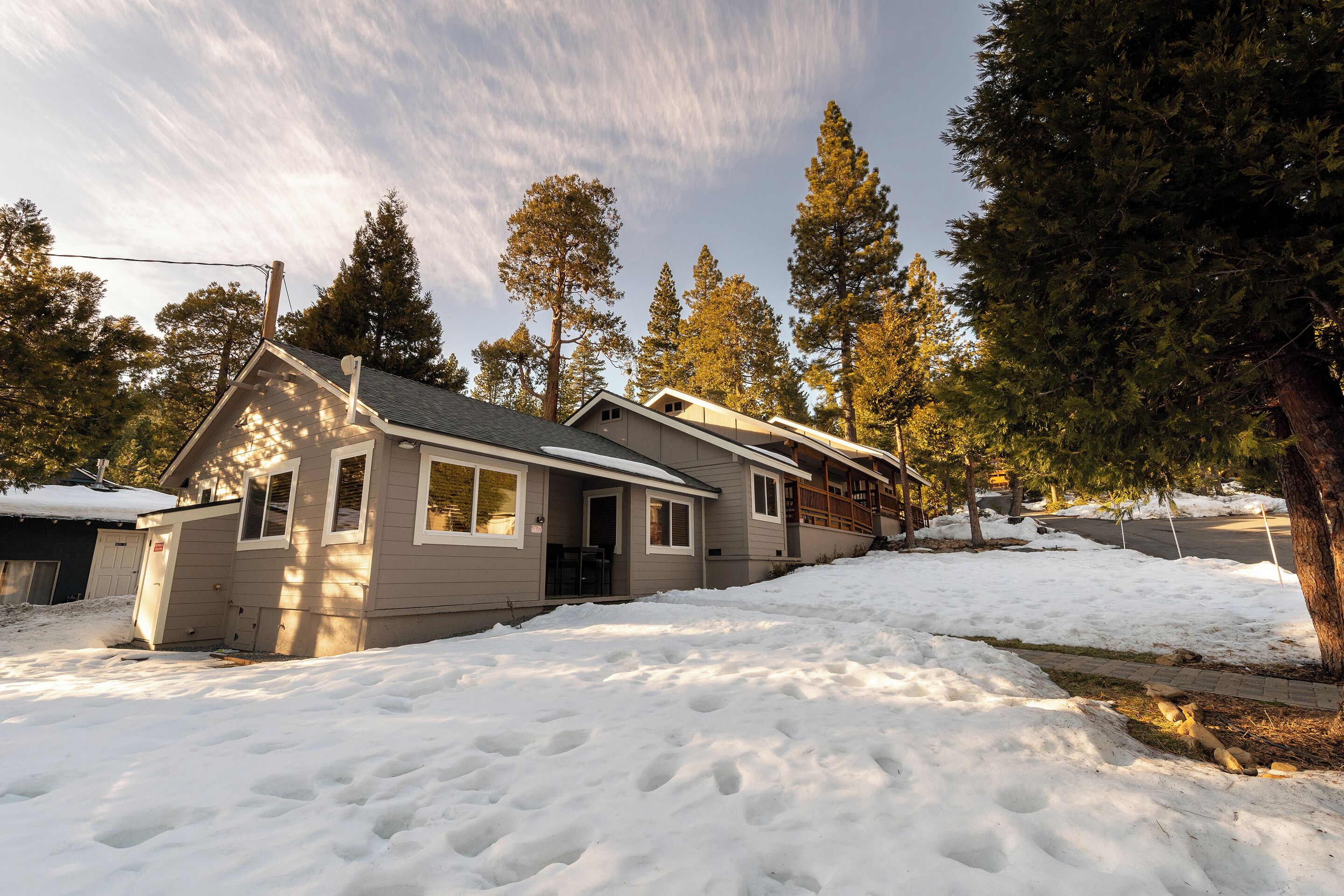 A two-story house with a covered porch and a garage, surrounded by snow and tall pine trees.