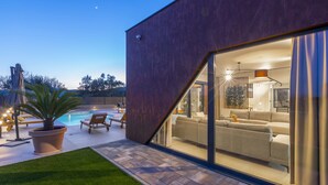 Una casa moderna con piscina, mobiliario exterior e interior bien iluminado visible a través de amplios ventanales.