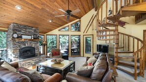 A spacious living room with a stone fireplace, leather sofas, a wooden ceiling fan, and a spiral staircase.