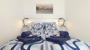 A neatly made bed with a patterned bedspread, two folded bathrobes on the bed, and a framed picture of a coastal town on the wall above the bed. - House of the Sun - VoudeFerias.pt (Albufeira)