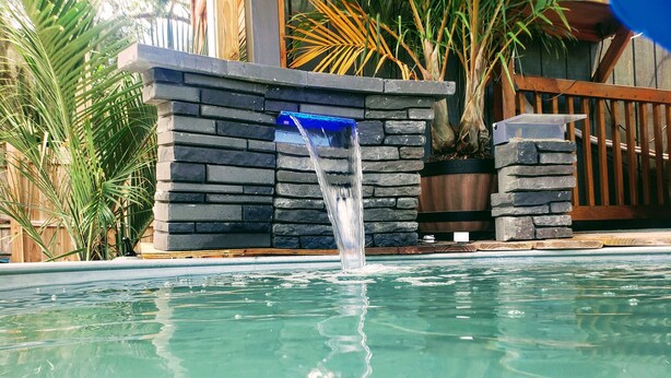 A swimming pool with a waterfall feature, surrounded by stone walls and tropical plants. - Tiny House , Big Vibes Illuminated Backyard Escape (St. Petersburg)