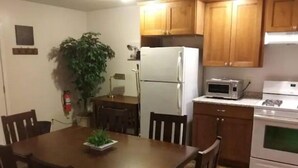 A compact kitchen with wooden cabinets, a white refrigerator, a microwave, and an oven. There is a dining table with chairs and a potted plant on it. - Lyle &Taylor Present -Slice of Home on the Road (Bourbonnais)