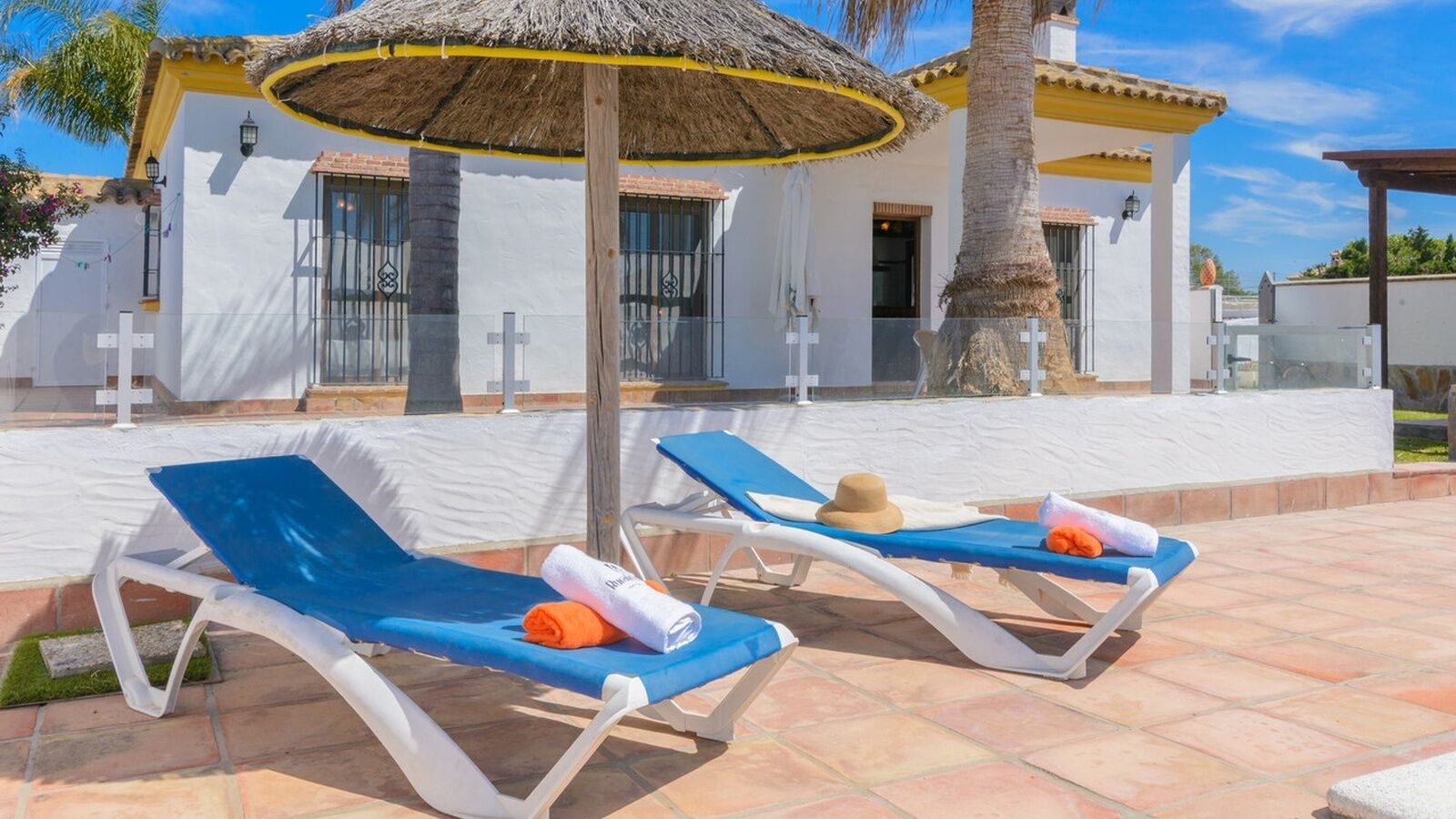 A pool area with two sun loungers, a thatched umbrella, and a white building with a yellow roof.