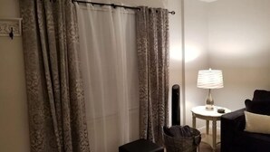 Una habitación con cortinas estampadas, una pequeña mesa con lámpara, una silla y un reposapiés negro.
