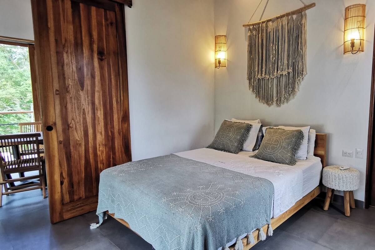 A bedroom with a wooden bed, a small stool, and a wall-mounted macrame light fixture.