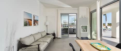 A modern living room with a sofa, a dining table, and a view of the kitchen and outdoor area.