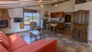 A cozy living area with a fireplace, a sofa, and a dining table with chairs. There's a kitchen with wooden cabinets and a large window offering a view of the outdoors. - Cortijo la Era Castril by Ruralidays (Granada)