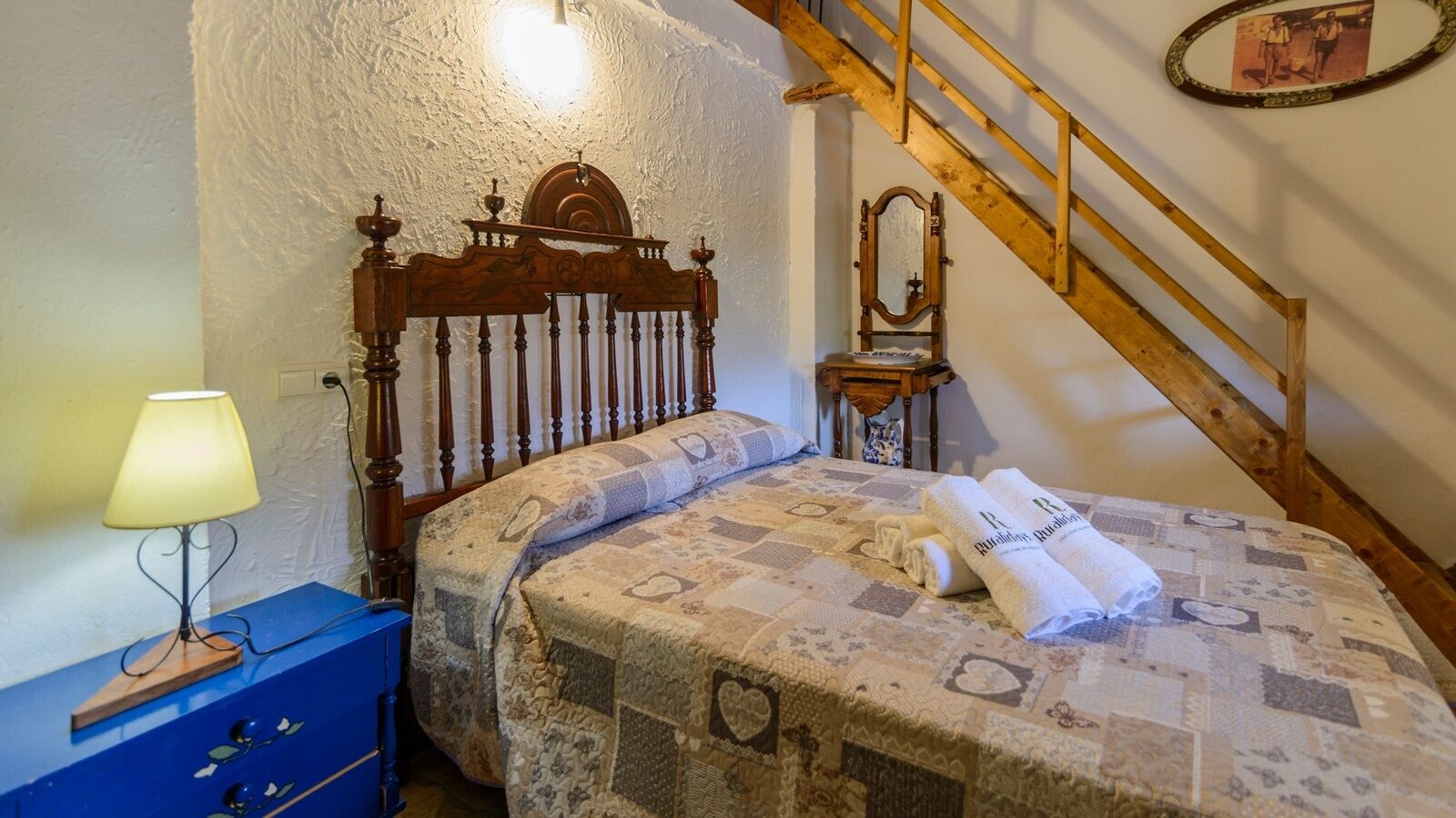 A bedroom with a wooden bed, a nightstand with a lamp, a mirror, and a staircase.