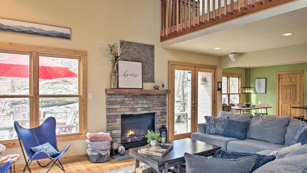 A cozy living room with a fireplace, a sofa, a coffee table, and a view of the outdoors through large windows. - Wintergreen Resort Cabin: Near the Slopes! (Wintergreen Resort)