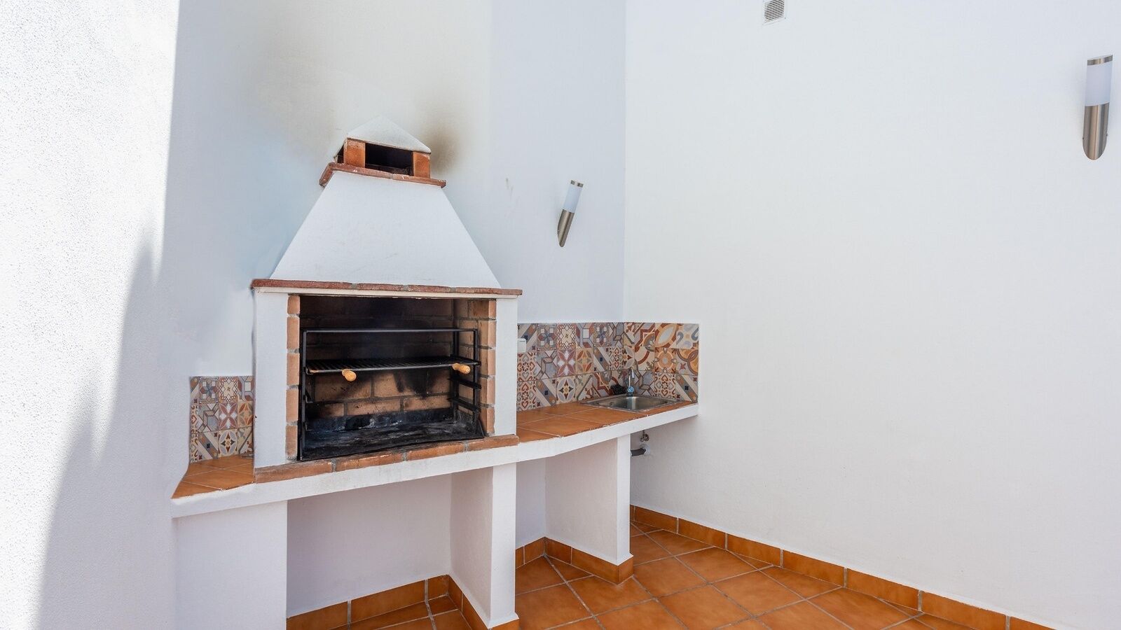 A small outdoor kitchen with a built-in grill, a tiled backsplash, and a white wall.