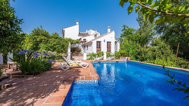 A swimming pool with a tiled edge, surrounded by a garden with various plants and a white building in the background. - Molino Maroto Alhaurín el Grande by Ruralidays (Málaga)