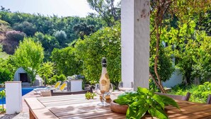 A wooden table with a bottle of wine, a glass, and a potted plant in a lush garden setting with a pool and lounge chairs. - Molino Maroto Alhaurín el Grande by Ruralidays (Málaga)