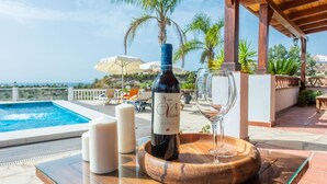 Une bouteille de vin sur un plateau en bois, accompagnée d’un verre, de bougies, et une scène se déroulant au bord d’une piscine, avec des parasols et des chaises longues.