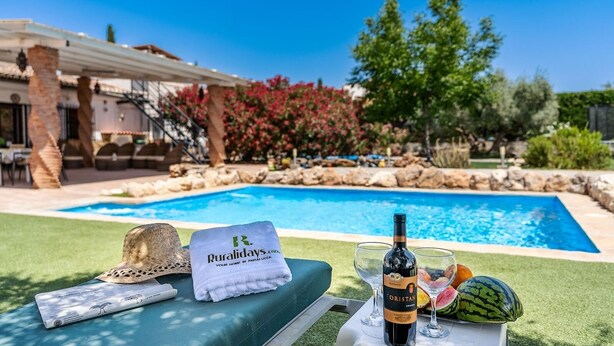 A poolside setting with a lounge chair, a towel, a bottle of wine, and a fruit bowl.