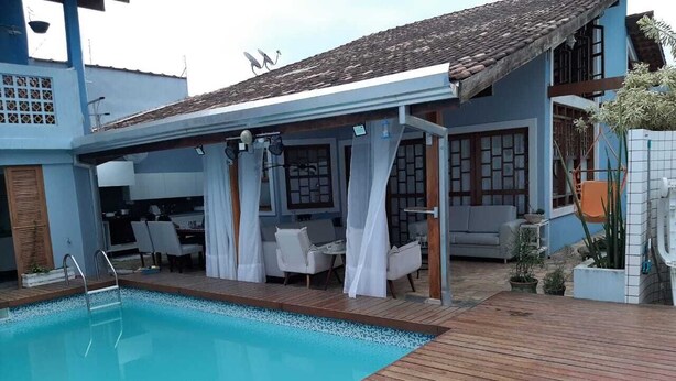 Una piscina con terraza de madera, una zona de estar techada con cortinas blancas, y un edificio con paredes azules y tejado de baldosas.