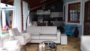 Un salón con un sofá blanco, una mesa de centro con adornos, una cocina al fondo y una zona de piscina visible a través de una puerta abierta.