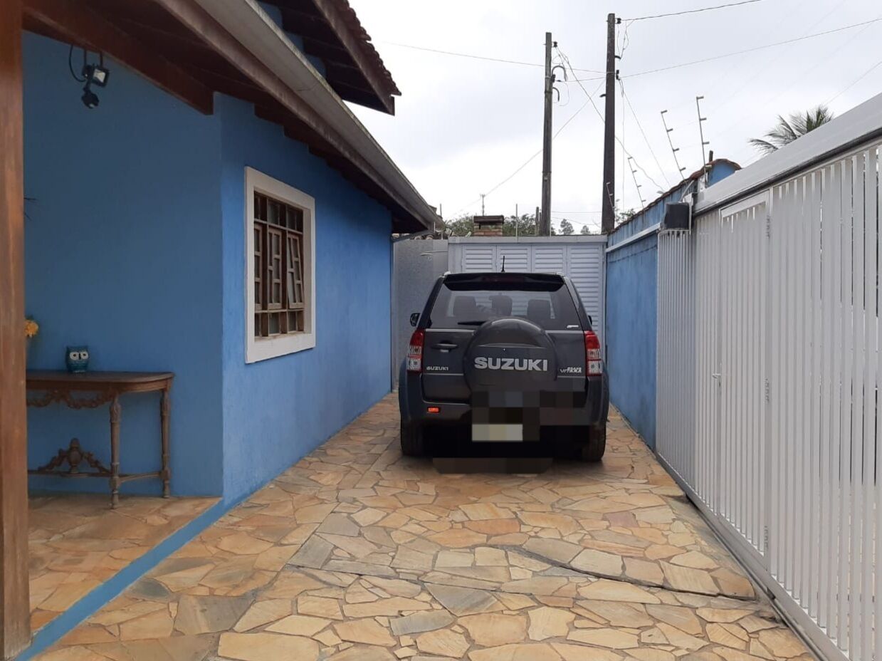 A narrow stone-paved alleyway with a parked Suzuki vehicle, a blue wall, and a white fence on one side.