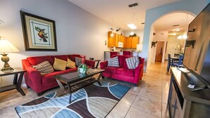 A living room with a red sofa, a coffee table, and a rug. There is a kitchen in the background with orange cabinets and a dining area with blue chairs. - Lovely 3BR 2 Bath Orlando Area Resort Condo (Davenport)