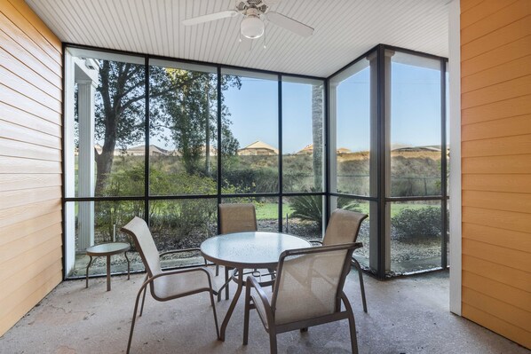 A glass-enclosed patio with a round table and four chairs, offering a view of a landscaped garden and distant hills.