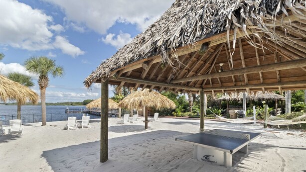 Un espace aménagé au bord de la plage, avec un pavillon à toit de chaume, des chaises longues blanches et une table de ping-pong.