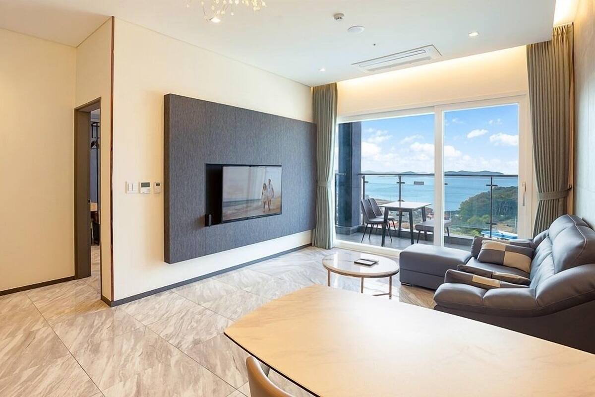 A modern hotel room with a large TV mounted on the wall, a sofa, a small table, and a balcony with a view of the sea.