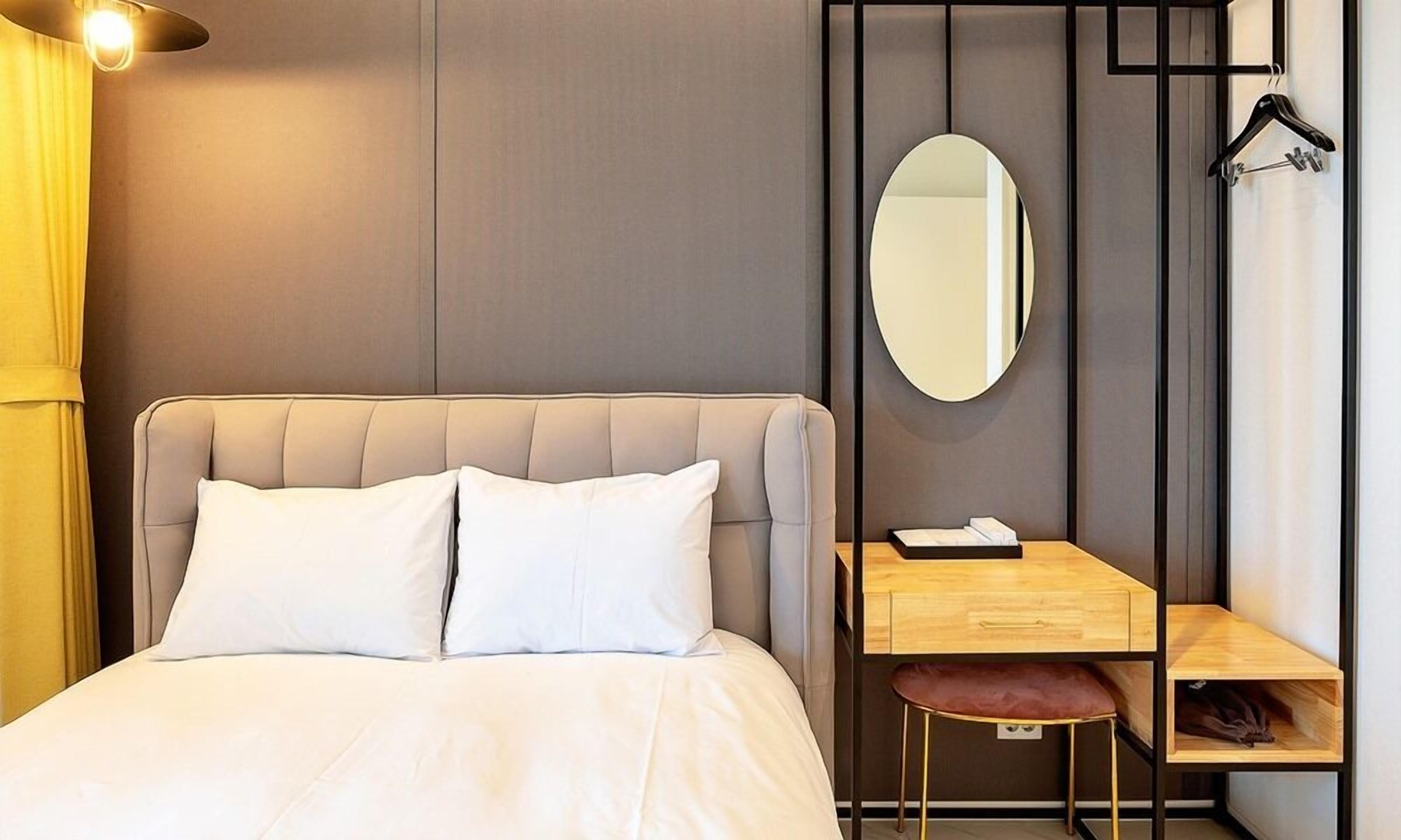 A modern hotel room with a large bed, a desk with a chair, a mirror, and a hanging coat rack.