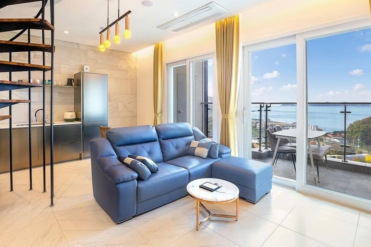 A modern living room with a blue sofa, a round coffee table, and a view of the sea through large windows.