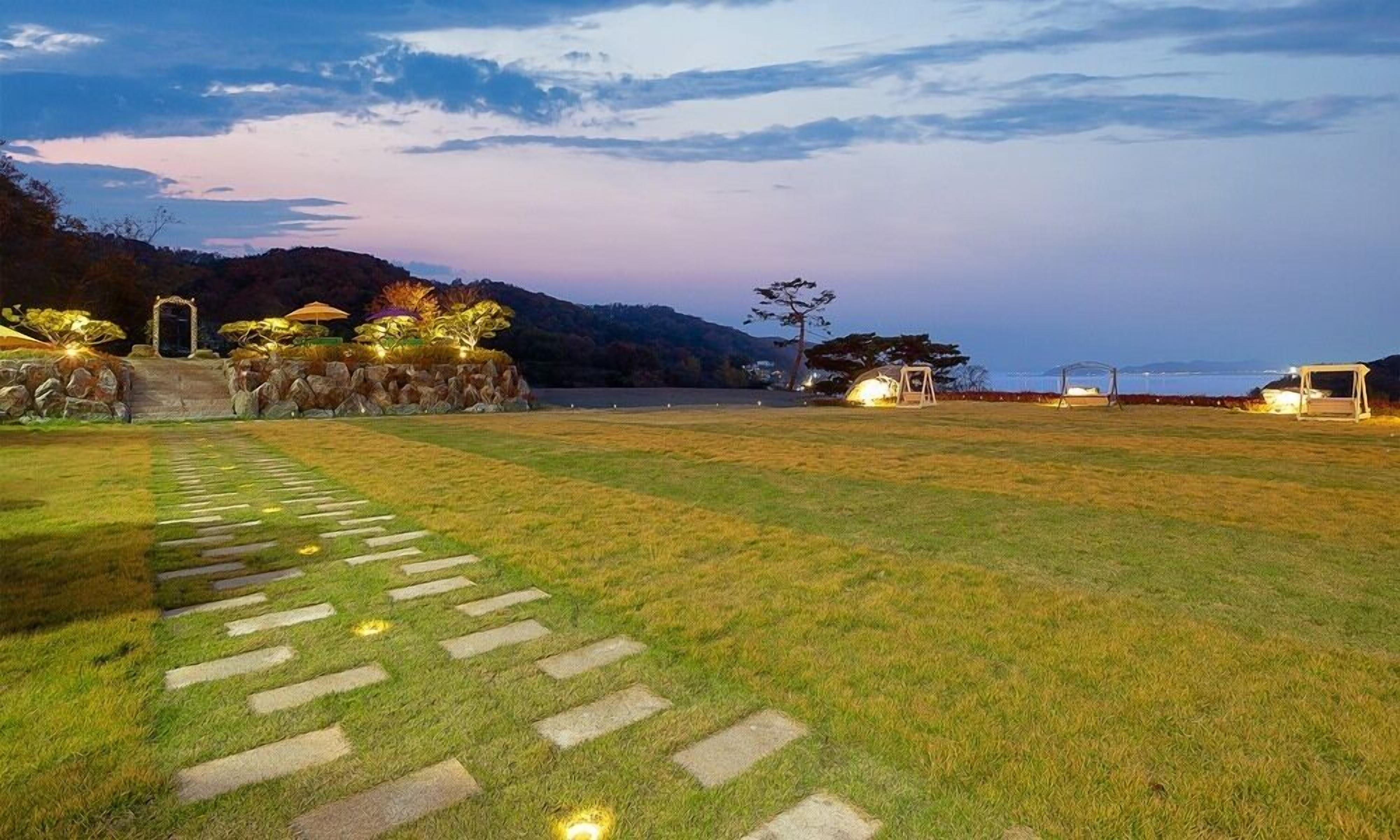 A well-maintained grassy area with stone pathways leading to a seating area with umbrellas, overlooking a body of water and hills.